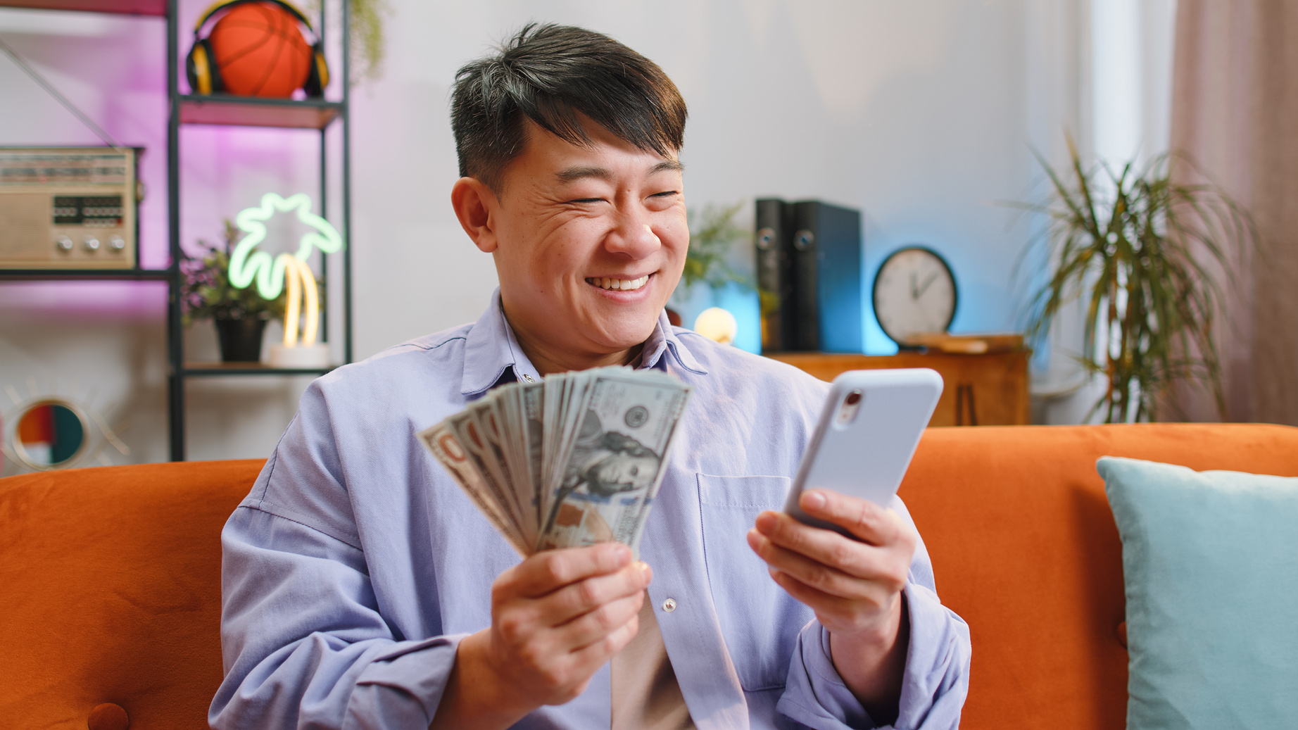 Smiling happy asian man counting money cash use smartphone, income saves lottery win, budget at home